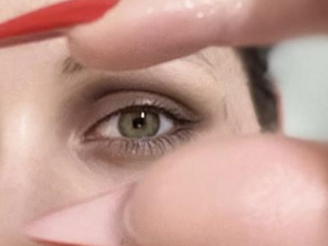 LexyClaire's green eye being gently touched by a red eyeliner brush, her eyelash slightly raised, focusing on the precise application. Her skin is smooth, brownish, and she's looking forward.