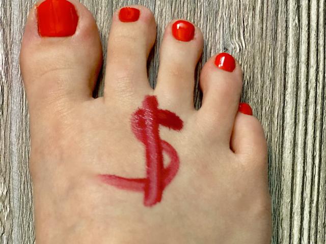 LexyClaire's foot with red painted nails, a red dollar sign tattoo on the sole, against a wooden surface. Her toes are slightly curled, showing the tattoo clearly.