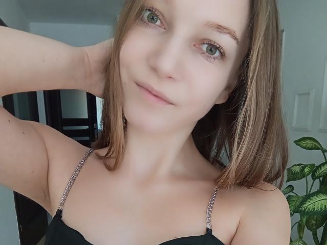 LovelyxEmilly with light brown hair, green eyes, and a slight smile, wearing a black strap dress with silver chains, has her arm behind her head.