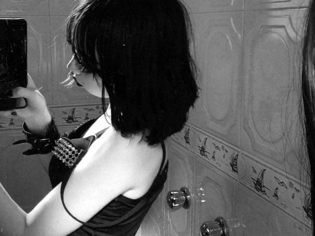 SilencePhsi with dark, wet, messy hair, holding a phone to her face, wearing a black, checkered bra, showing her bare, small, pale-ass in a black, sheer top.