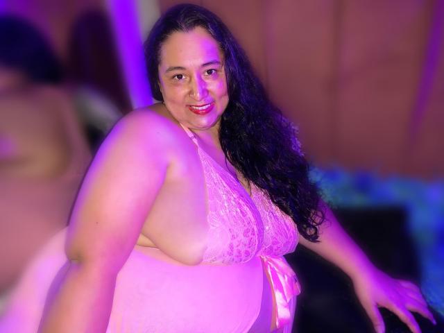TistMature has large, round tits in a pink lace bra, smiling with long curly black hair, pink and purple lighting, and a slightly blurred background.