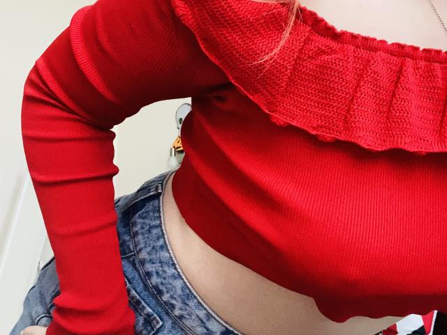 KarKutie wearing a red ribbed off-shoulder top, exposing her midriff and blue jeans. Her red top highlights her tits, and her hand rests on her hip.