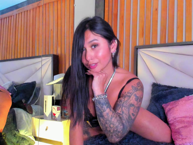SarahOrtiz, laying on a bed, has long black hair, tattooed arm, and is wearing black lingerie, with her hand on her chin, looking at the camera.