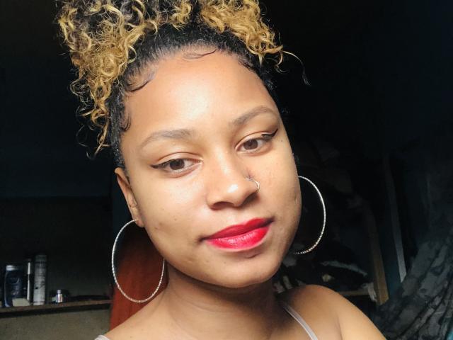 Mickaia with curly honey-brown hair, brown eyes, red lipstick, nose ring, and large hoop earrings, smiling softly. Her tan skin is smooth, and she's wearing a thin strap top.