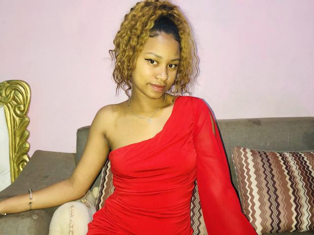 Mickaia in a red one-shoulder dress, curly brown hair, sitting on a gray couch with a zigzag-patterned pillow, looking at the camera with a slight smile.