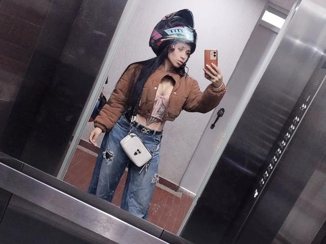 EvaMueller, com cabelos pretos longos, jaqueta marrom e volumosa, jeans rasgados, bolsa transversal branca e capacete, tirando uma selfie no espelho dentro de um elevador, mostrando seus seios pequenos..