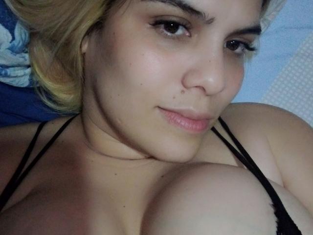 Febe Saron mit heller Haut, blonden Haaren und schwarzer Spitze, die in einem Selfie ihre großen Brüste zeigt, mit leicht geschürzten Lippen, die in die Kamera lächelt..