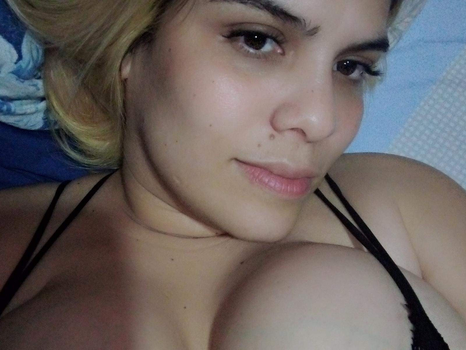 FebeSaron, com pele clara, cabelo loiro e lingerie preta, exibindo seus seios grandes em uma selfie, lábios levemente curvados, olhando para a câmera com um sorriso discreto..