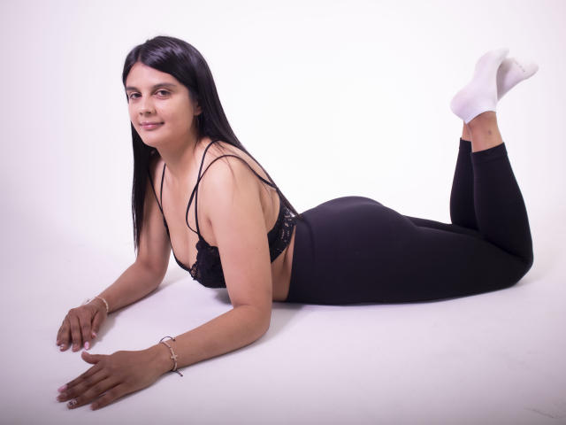 TiffanyGreaty is lying on her stomach with black lace bra, black leggings, and white socks, smiling at the camera. She has long black hair and a cute butt.