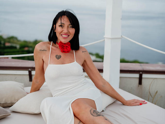 AphroditeG mit schwarzen Haaren, die ein weißes Kleid trägt, eine rote Blumenkette um den Hals hat, Tätowierungen auf Brust und Oberschenkel, sitzt auf einem Bett, lächelt, im Hintergrund ist das Meer zu sehen, weiße Säulen..