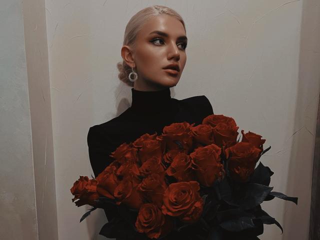 SelenaeMoore, avec des cheveux blonds platine, un pull noir à col roulé, tenant un bouquet de roses rouges, regardant sur le côté avec une expression sérieuse, portant de grandes boucles d'oreilles en argent..