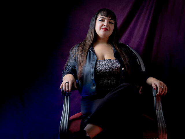 CherryMeloon sits on a chair with black arms, wearing a black leather jacket, leopard print top, and black pants, her red lipstick contrasting with her dark hair.