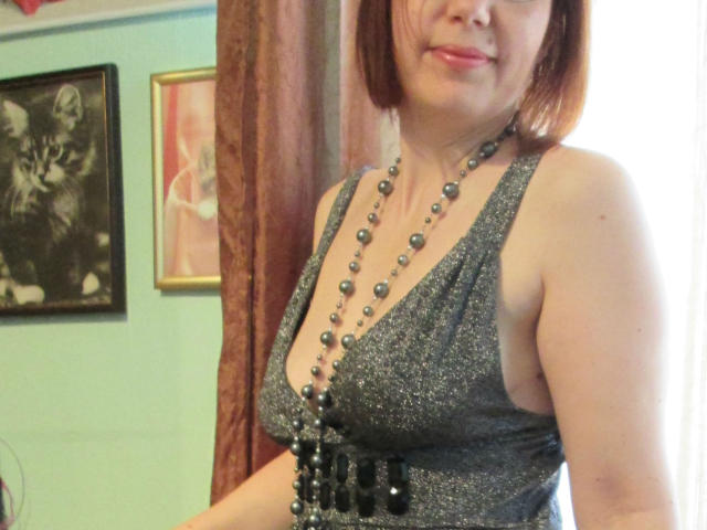 JuicyLady69 with shoulder-length brown hair, wearing a sparkly grey dress with a deep V-neck, black beaded necklace, and black beaded belt, smiling slightly. Boobs visible.