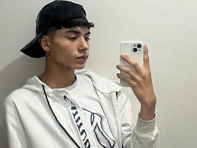 ImJuanda with short black hair, wearing a black baseball cap, white hoodie, and taking a selfie with a white iPhone. Small facial piercings visible. Serious expression.