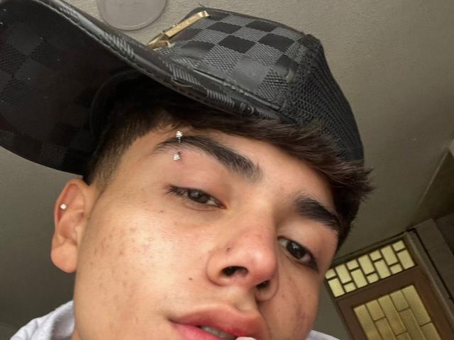 ImJuanda has a nose piercing, a black checkered hat, a single ear piercing, and a small nose stud, with short brown hair and a slightly flushed face.