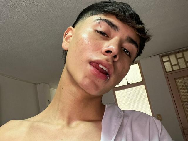 ImJuanda has a shiny face, short black hair, and a nose ring. He's sticking out his tongue with slightly parted lips, wearing a white shirt, and looking down.