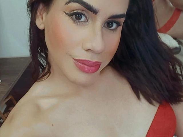 KamilaGrant has red lipstick, dark eyebrows, and black hair. She's wearing a red top, showing her bare shoulders and cleavage. Her tits are small to medium. She's smiling.
