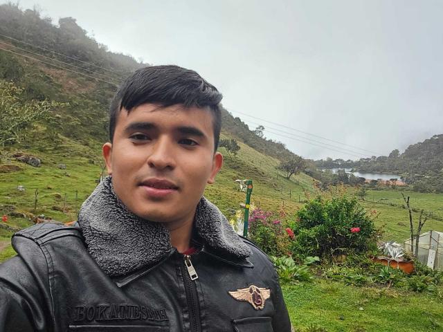 DayanWilly, mit kurzen, schwarzen Haaren und einem leichten Lächeln, trägt eine schwarze Lederjacke mit einem grauen Pelzkragen und steht in einer grünen, hügeligen, nebelverhangenen Landschaft..