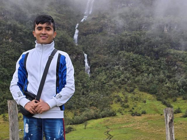 DayanWilly with short black hair, white and blue jacket, jeans, black strap, standing in front, hands together, smiling, waterfall in foggy green forest behind.