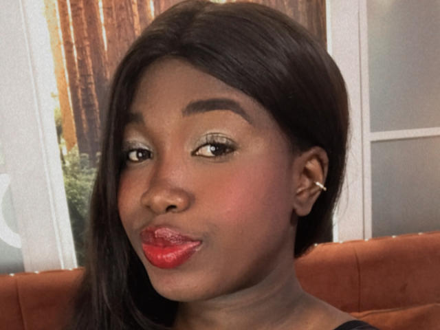KarlieMoss has dark brown skin, long black hair, and is wearing red lipstick. She has subtle eyeshadow, a small nose, and a cute, slightly pouty face with a small smile.