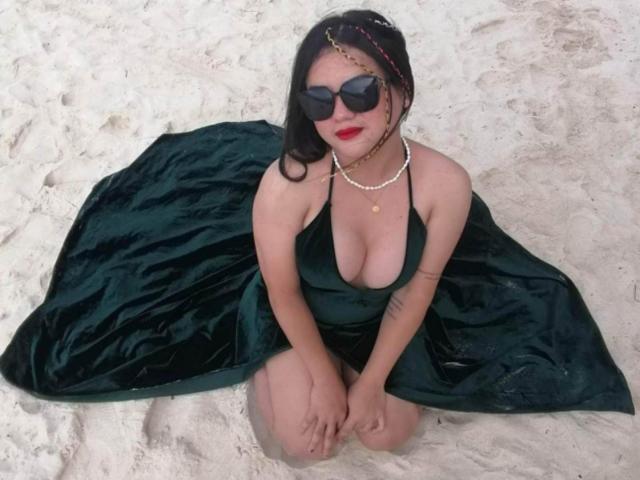 YourSophie, con i capelli scuri e il rossetto rosso, è inginocchiata sulla sabbia, indossa un abito di velluto verde, occhiali da sole neri e una collana di perle, mostrando il seno in modo carino e sensuale..