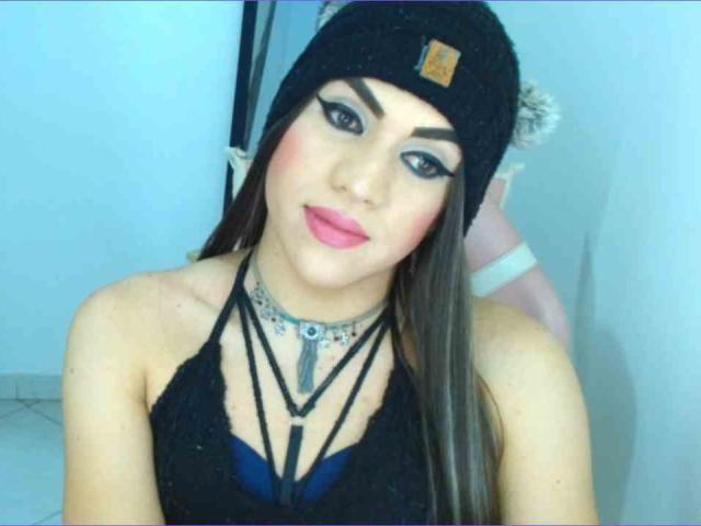 GeishaSenier with long brown hair, black beanie, bold eyeliner, pink lipstick, black strappy top, choker, and necklace, showing her tits, in a selfie.