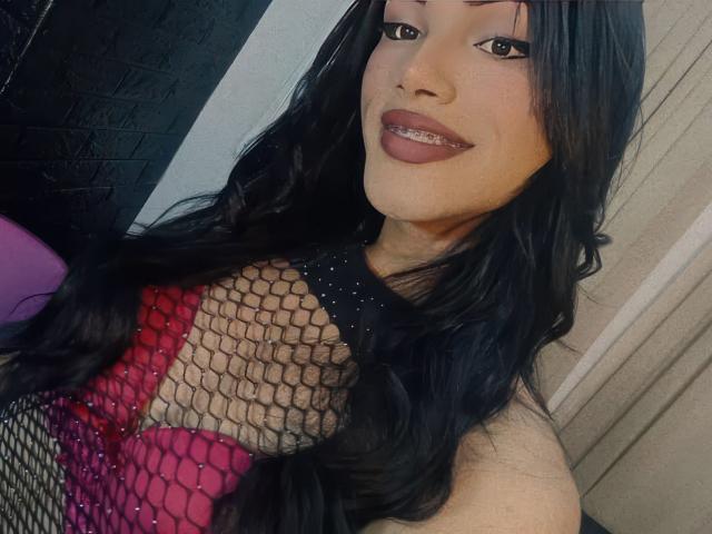 KittyQueeny with long black hair, wearing a fishnet top over red and pink sequined fabric, showing her tits, smiling with brown eyes and pink lipstick.