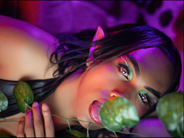 KittyQueeny with dark wet hair, green eyelids, and pink lips, holding green leaves, her tongue slightly out, purple and pink lights on her face, elf ears visible.