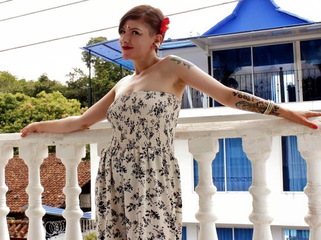 MaferCougar with short brown hair, red flower in hair, tattooed arm, strapless floral dress, leaning on white balcony, blue-roofed building in background, standing with arms out.