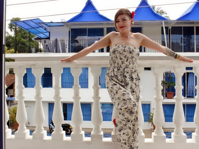 MaferCougar with short brown hair, red flower in hair, strapless floral dress, arms on white balcony, standing, tattoo on right arm, smiling, blue roofed building in background.
