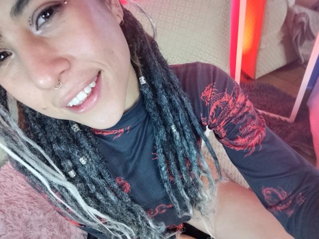 HellenK with black and white dreadlocks, nose ring, smiling, wearing a black shirt with red dragon design, white teeth, pink lips, and a cute face. 