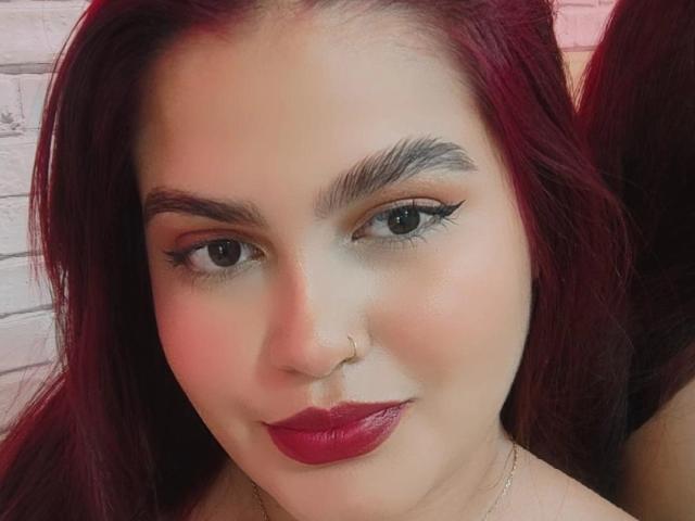 AbbyLester has red lipstick, thick black eyebrows, and long red hair with a nose ring. She's smiling with dark eyeliner and a gold necklace.