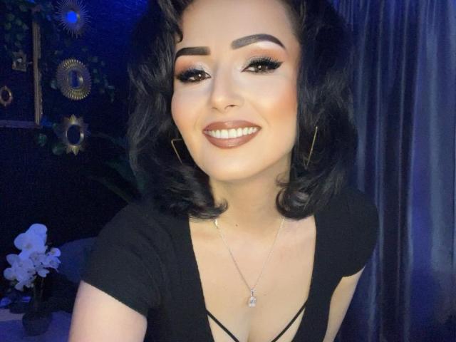 MazikeenMaze with curly black hair, thick eyebrows, dark lipstick, and a smile, wearing a black top with a low neckline, showing her tits. She's wearing gold earrings and a necklace.