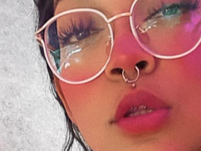 DulceTammara69's face with pink-tinted skin, pink glasses, nose ring, and pink lips, wearing green eyeshadow, looking up with slightly parted pink lips.