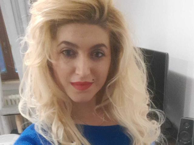 Blonde ElizabethRaegan with wavy hair, red lipstick, and blue sweater, smiling slightly. Boobless, no visible tits or pussy. Face is centered, hair is voluminous. Bedroom background.