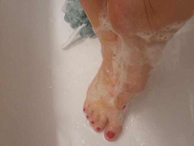 ElizabethRaegan's soapy foot with red-painted toes in a white bathtub, showing her toes and slightly curled pinkish toenails, with bubbles covering parts of her foot.