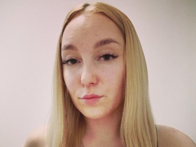 Dina Toni ha lunghi capelli biondi, lentiggini e sopracciglia folte con lunghe ciglia, indossa un'espressione neutra ed è rivolta leggermente di lato con un sorriso appena accennato..