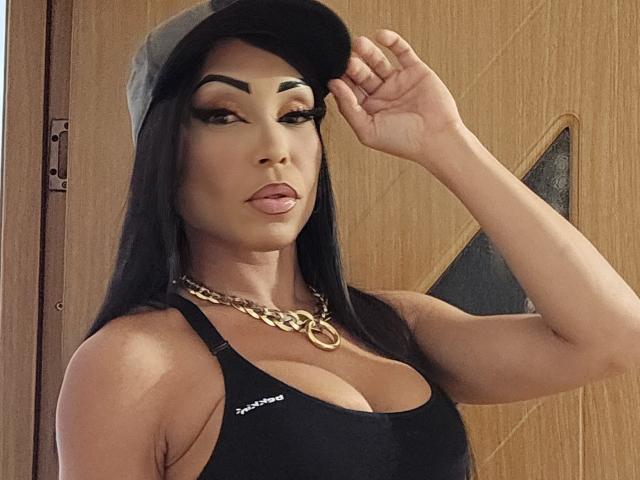 SquirtLatinQueen with long black hair, wearing a black cap, black tank top showing big boobs, gold chain necklace, and makeup, touching cap, looking at camera.