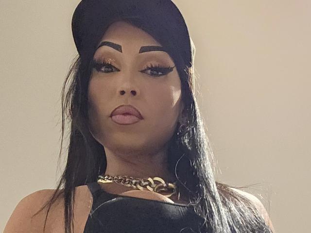 SquirtLatinQueen has dark eyebrows, long black hair, a black cap, gold chain, and a black top, with her tongue slightly out, looking serious and sexy.