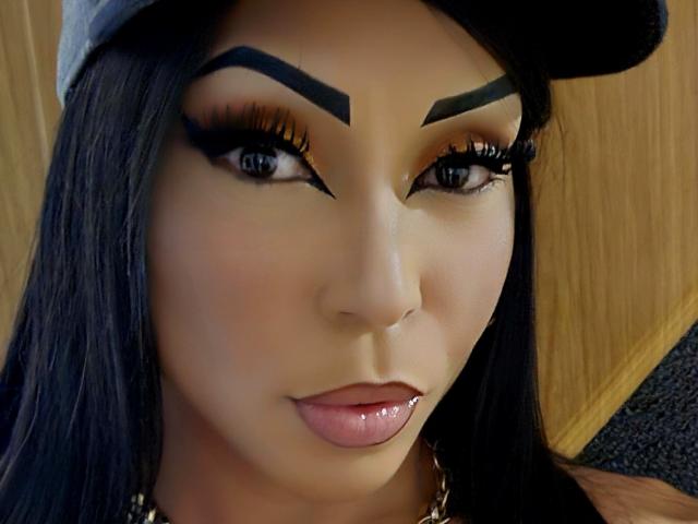 SquirtLatinQueen has thick, black eyebrows, long, dark eyelashes, and glossy pink lips. She's wearing a gray cap and has long black hair. Her skin is smooth and tan.