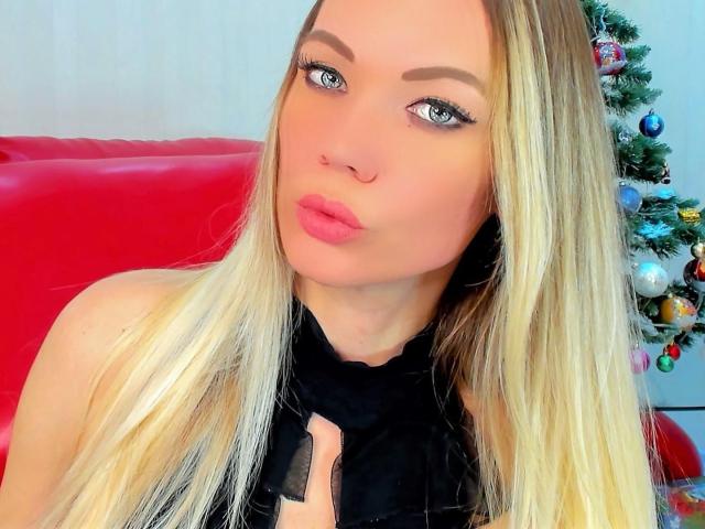JuliaLoves has blonde hair, blue eyes, and pink lips. She's wearing a black top, with a red chair and decorated Christmas tree in the background.