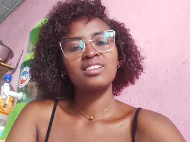 Lalance has dark brown skin, curly black hair, wearing clear glasses, black spaghetti strap top, gold necklace, and a nose ring. She's smiling with shiny lips.