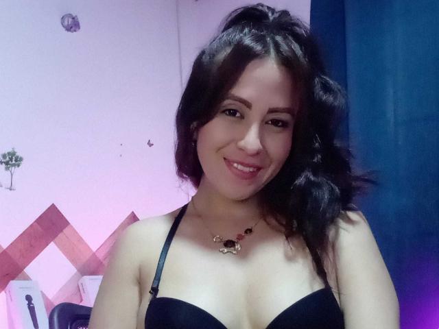 CamileParist with black hair, smiling, wearing a black bra, showing her tits, has a necklace with a small animal charm, in a room with blue and pink walls.