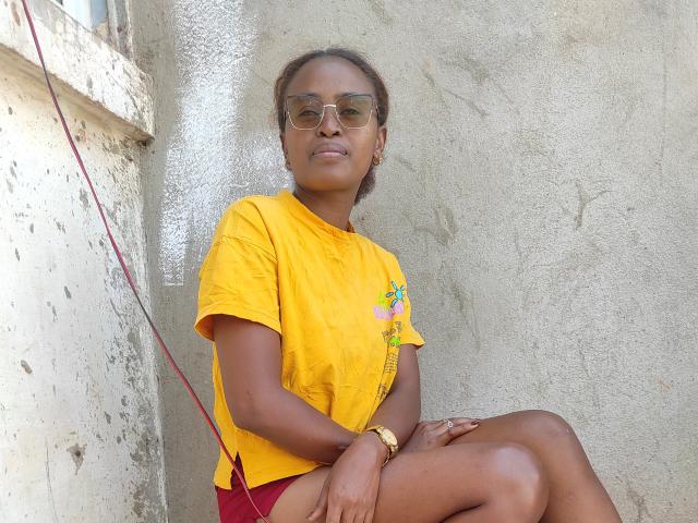 Lalance with dark brown skin, wearing a yellow tee and red shorts, has small tits, a gold bracelet, and green sunglasses, sitting against a textured wall.