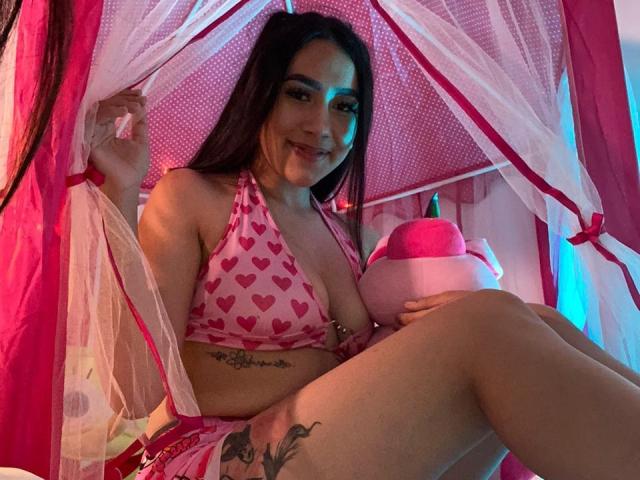 CelesteRusso with long black hair, pink heart-patterned bikini, and tattoos, smiling, holding a pink stuffed toy, sitting in a pink and white canopy bed, showing her tits.