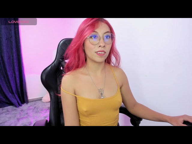 NattOne with red hair, glasses, and a yellow tank top, sitting in a black chair. Her tits are slightly visible through the thin top. She's smiling with pink lipstick.