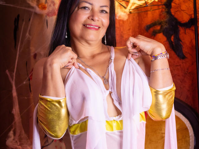 AnaMaturee with dark hair, smiling, pulling up her sheer pink dress, revealing her small tits and a yellow belt, wearing gold arm cuffs, and a silver bracelet.