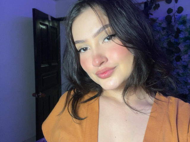 NatiQueen has medium-length black hair, light skin, and a subtle smile with pink lips. She's wearing an orange top, showing her small tits. Blue light in the background.