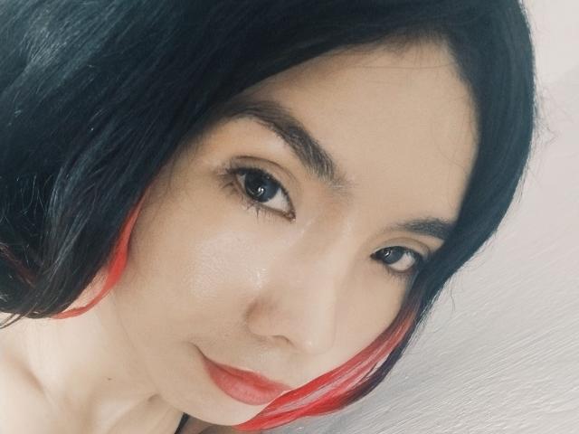 MileyWatson with black hair, red highlights, and a subtle smile, has smooth, fair skin and red lipstick, looking at the camera with dark, arched eyebrows.