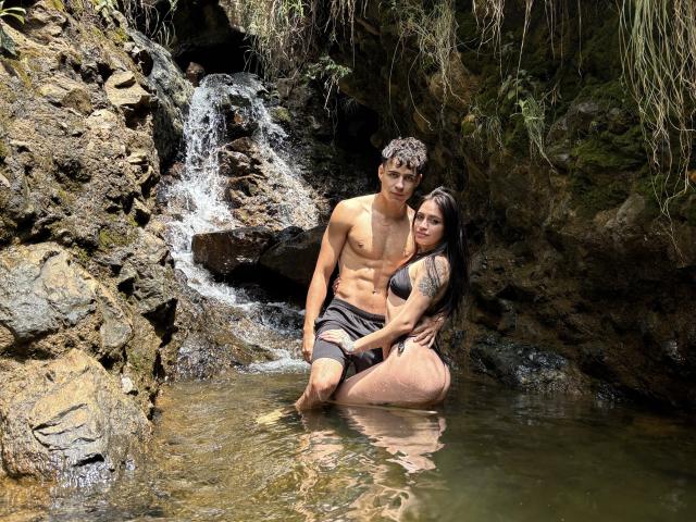 SofiaAndgeordi with curly brown hair, shirtless, wearing black shorts, kneeling in water. SofiaAndgeordi with long black hair, tattoo on arm, in black dress, touching SofiaAndgeordi's ass.